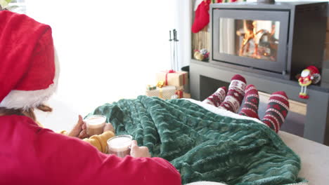 Diverse-gay-male-couple-drinking-cocoa-on-sofa-by-fireplace-at-home-at-christmas,-slow-motion