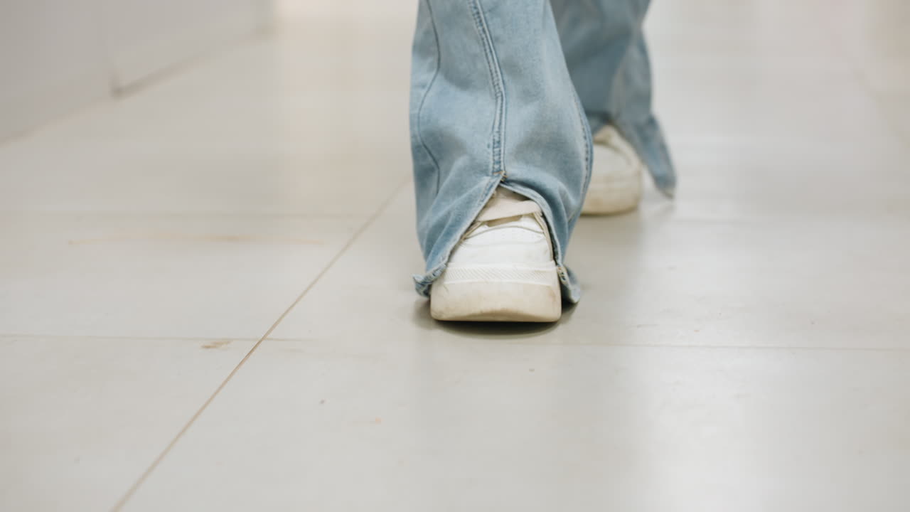 Close view fabric care professional legs in jeans and white sneakers walking along bright laundromat corridor, focused steps on tile floor, casual indoor mood with curtain, modern clean ambiance