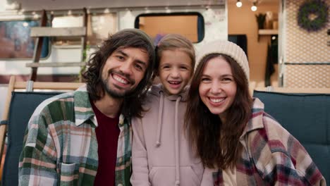 Portrait-of-a-happy-family,-a-brunette-guy-with-stubble-in-a-Green-plaid-shirt,-a-brunette-woman-in-a-plaid-shirt-and-their-little-daughter,-a-blonde-girl-in-a-pink-hoodie,-standing-near-their-camp-trailer,-smiling-and-looking-at-the-camera-during-their-picnic-outside-the-city-in-the-summer