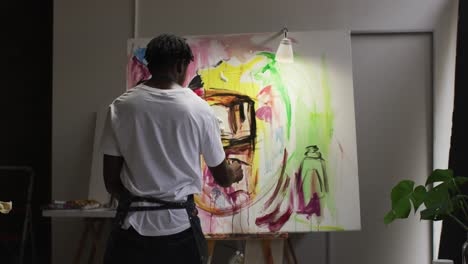 Rear-view-of-african-american-male-artist-wearing-apron-painting-on-canvas-at-art-studio