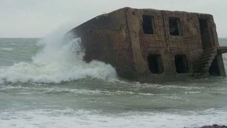 Big-stormy-waves-breaking-against-abandoned-seaside-fortification-building-ruins-at-Karosta-Northern-Forts-in-Liepaja,-Baltic-sea-coastline,-wave-splash,-rainy-overcast-day,-medium-shot