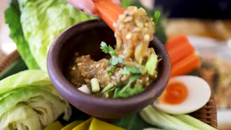 a hand dips a carrot stick into roasted green chili dip, surrounded by fresh vegetables and boiled egg, in bright natural light