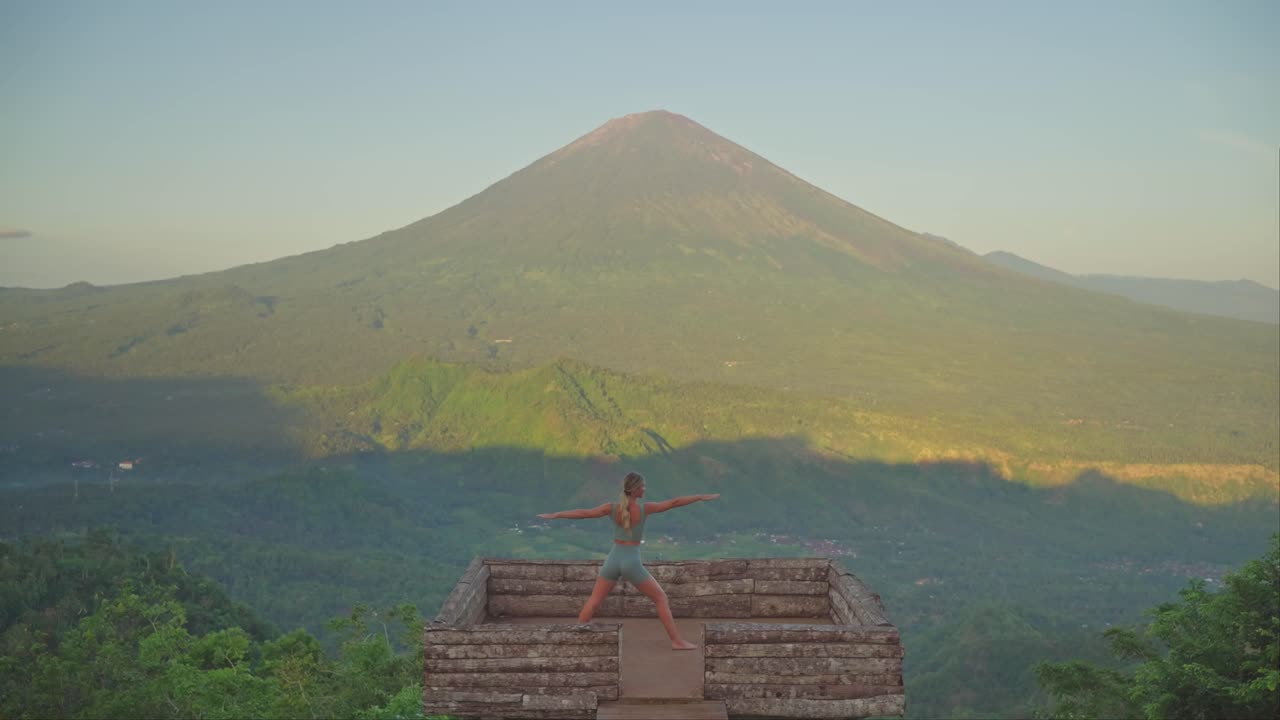 mujer practicando una postura de guerrera feroz en una plataforma de madera frente al monte agung