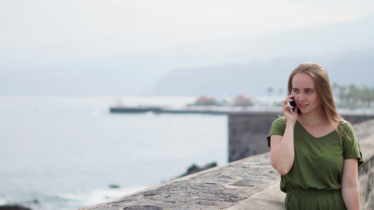 hermosa chica en un vestido verde hablando por teléfono de pie en la orilla cerca del océano y sonriendo
