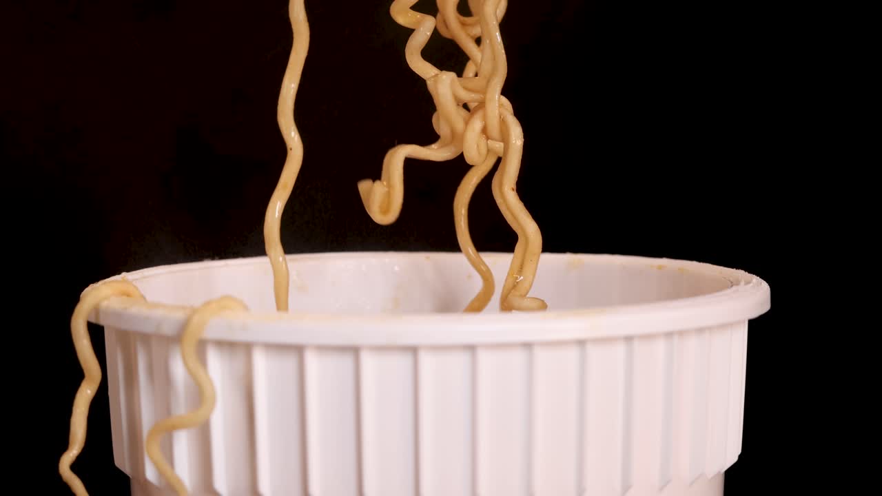 Una mano rilascia noodles istantanei cotti dalle bacchette in una tazza bianca su uno sfondo scuro, con inquadratura ravvicinata e illuminazione da studio uniforme.