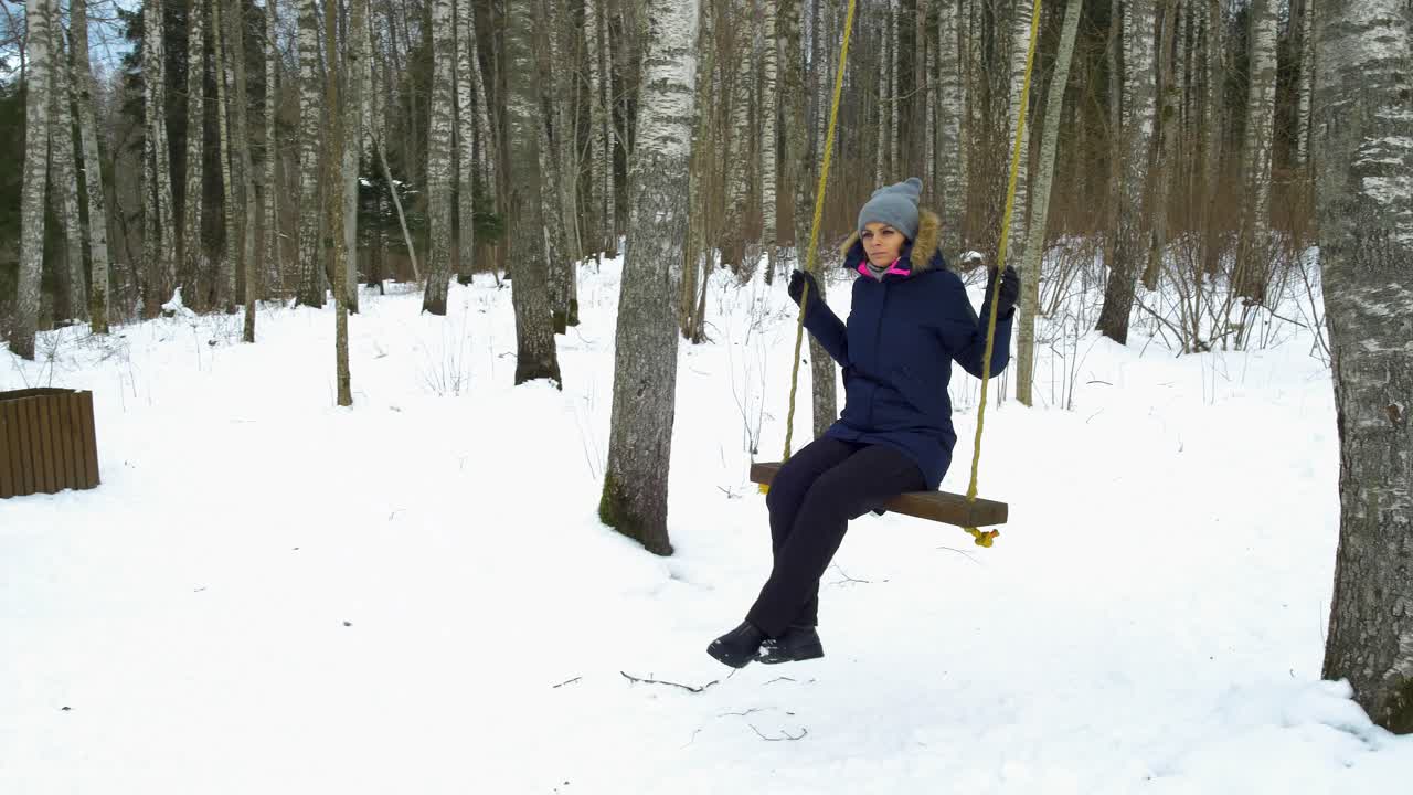 una joven caucásica balanceándose en el bosque de invierno en un bosque de abedules de invierno cubierto de nieve en un día soleado, sola en un clima helado, girando hacia el tiro de cardán derecho