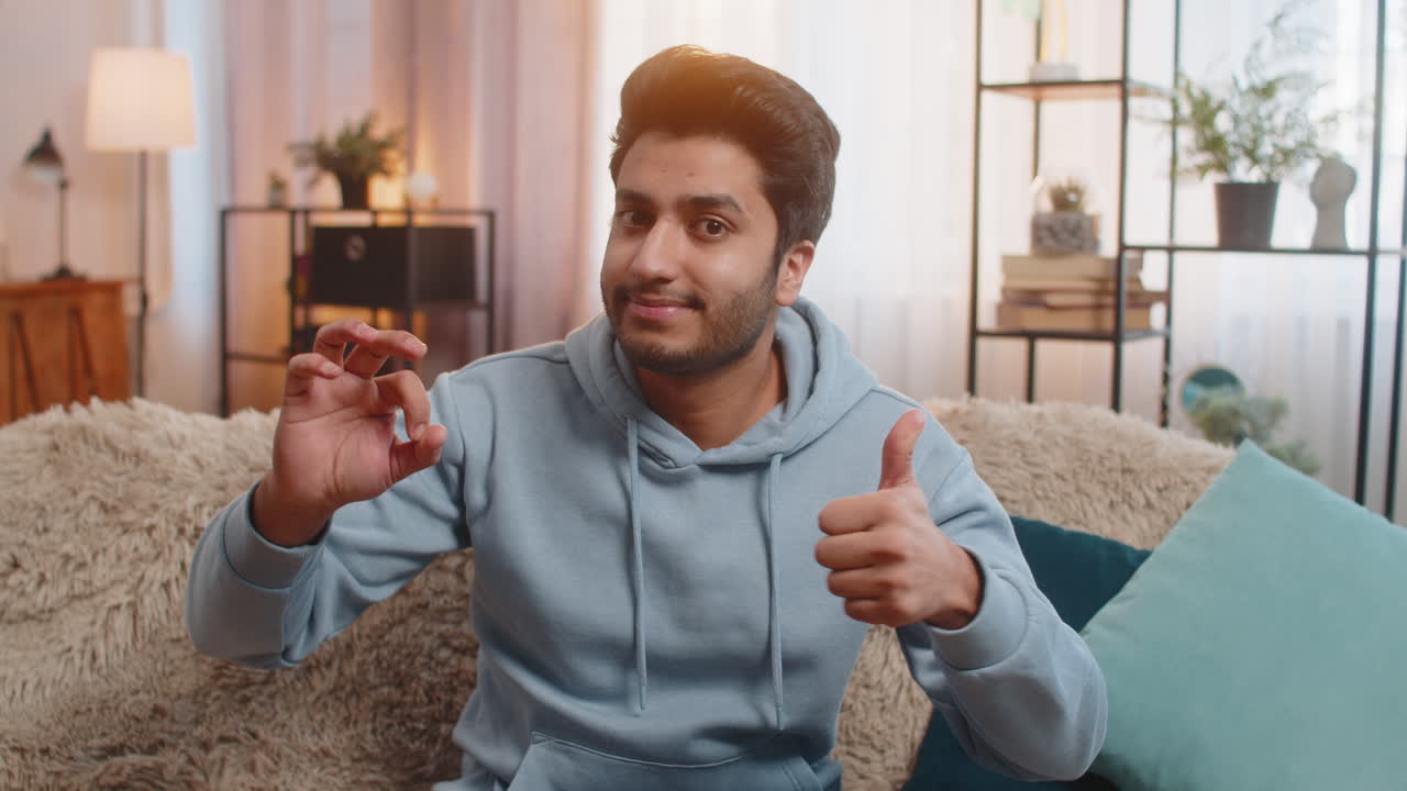 Happy indian man showing ok gesture and smiling confidently while sitting on sofa at home room