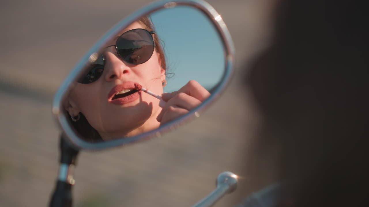 Primo piano di una donna riflessa negli occhiali da sole mentre si mette il rossetto usando lo specchietto laterale di una moto, che mette in risalto la bellezza, i dettagli e lo stile di vita all'aria aperta con la luce naturale del sole