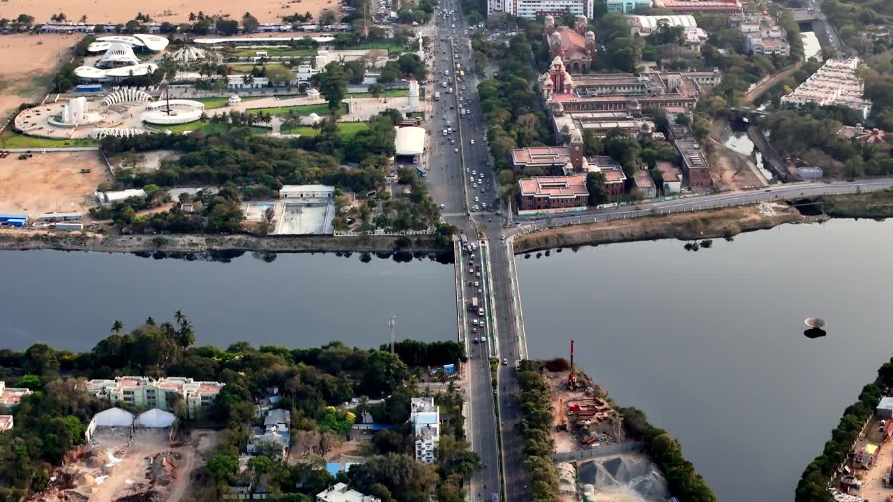 Ripresa drone direttamente sopra il Ponte Napier, che cattura veicoli che scorrono sulla strada simbolo. Il fiume Cooum scuro fiancheggia il ponte, creando un sorprendente contrasto tra il movimento urbano e l'immobilità.