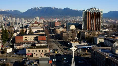 Flying-Above-The-Main-Street-And-E-12th-Avenue-Intersection-In-Vancouver,-BC,-Canada-With-View-Of-Landmarks-And-Mountain-Landscape-At-Daytime