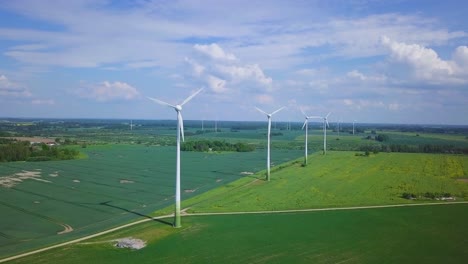 Aerial-view-of-wind-turbines-generating-renewable-energy-in-the-wind-farm,-sunny-summer-day,-lush-green-agricultural-cereal-fields,-countryside-roads,-wide-angle-drone-shot-moving-backward