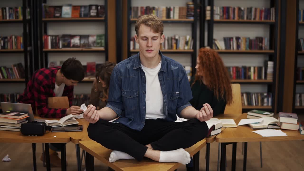 en la biblioteca durante el estudio. joven estudiante meditando en el escritorio en la pose del loto mientras sus compañeros de clase en el fondo continúan estudiando ignorándolo. estanterías en el fondo