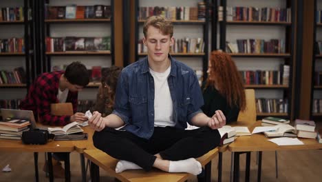 in the library during the studying. young male student meditating on the desk in the lotos pose while his classmates on the background continuing studying ignoring him. bookshelves on the background