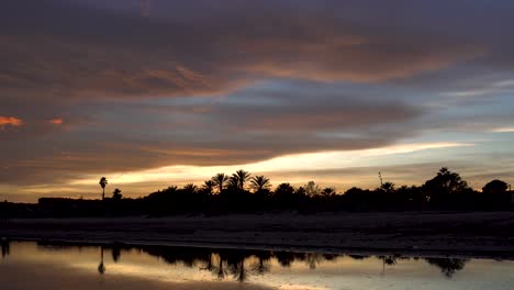 Zeitraffer,-Goldener-Sonnenuntergang-Mit-Orangefarbenen-Wolken,-Palmen-Und-Der-Silhouette-Zweier-Pferde,-Die-Im-Dunklen-Gegenlicht-über-Dem-Meerwasser-Am-Strand-Entlang-Spazieren