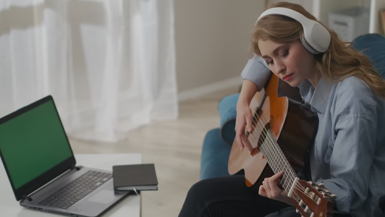 self-education of playing guitar by internet, young woman is playing song sitting in front of laptop with green screen
