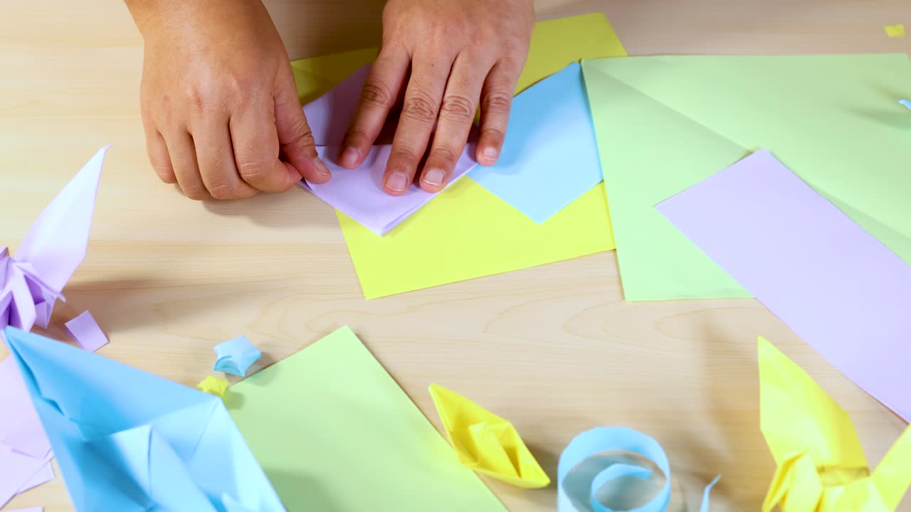 Person folds pastel origami paper, surrounded by finished crafts, under bright, even lighting overhead