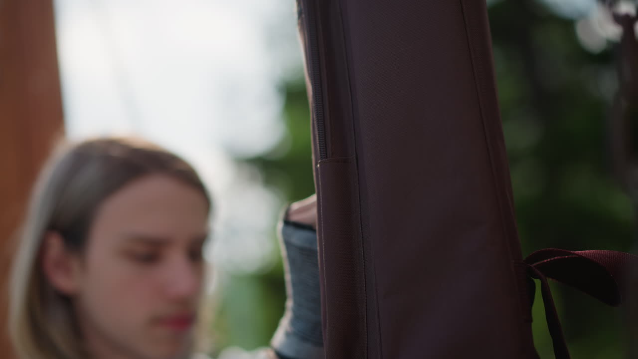 closeup teen guitarist holding brown case outdoors longhair musician opens zipper, adjusts strap, focused expression, suburban park background, natural light, candid rehearsal vibe, preparation