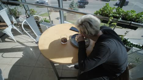 young man with blond hair enjoying a sandwich and iced coffee at a cafe table by a window with city views, natural light, and potted plants. relaxed and casual dining scene