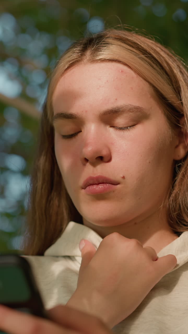 primer plano de una joven con una pequeña mancha facial usando el teléfono atentamente mientras sostiene su collar de capucha, la luz del sol iluminando suavemente su cara a través de las hojas de los árboles, con un fondo verde borroso