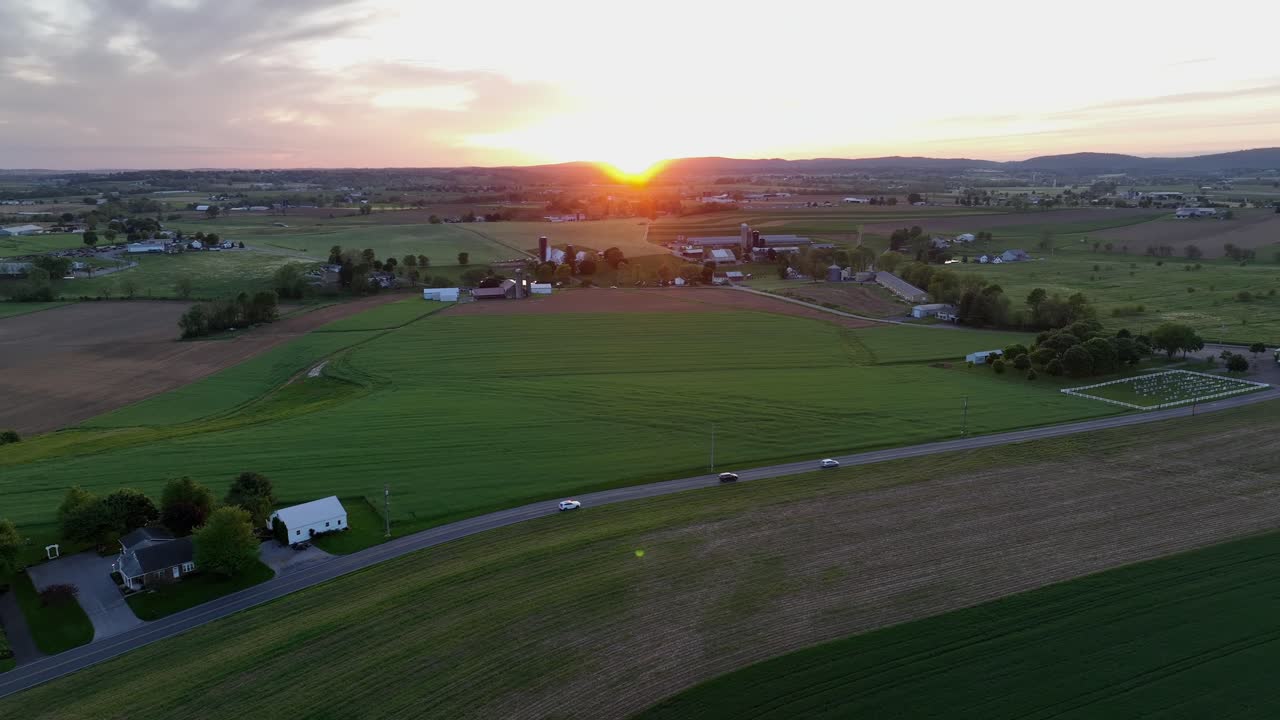 Cars on rural road of american suburb town at golden sunset. Aerial backwards wide shot. Agricultural fields and farm houses in Pennsylvania, USA