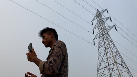 looking up at young indian male making call on smartphone under power line pylon