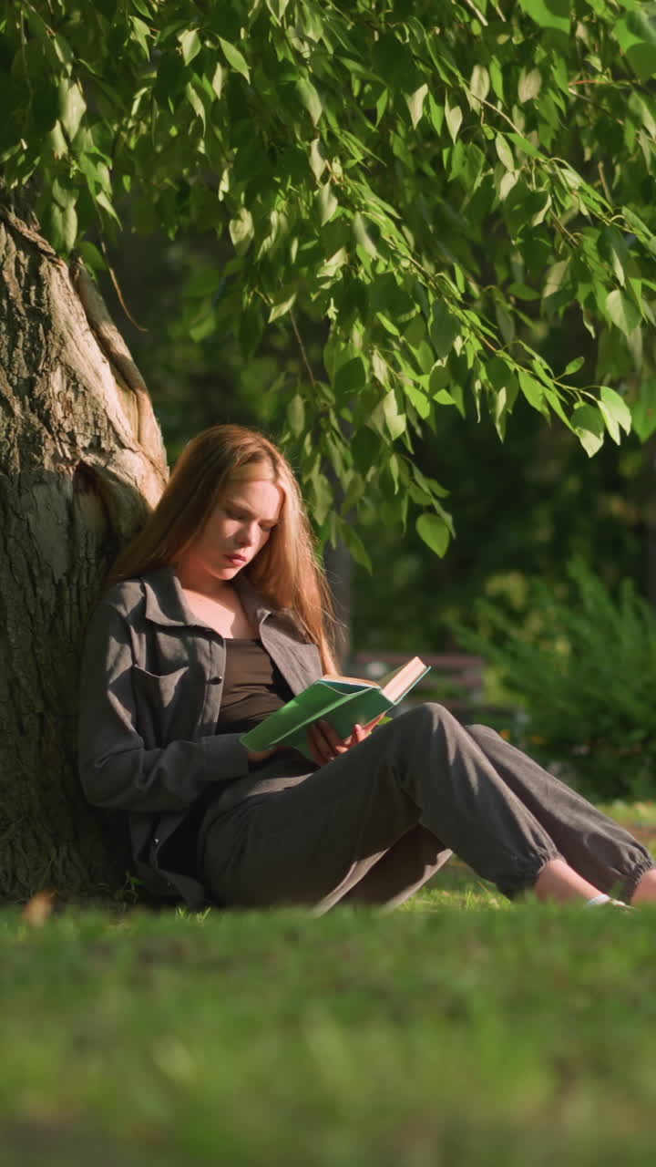 mujer sentada al aire libre, apoyada en un árbol en un campo de hierba, leyendo un libro bajo la cálida luz del sol, las hojas de los árboles se balancean suavemente en la brisa, el fondo muestra un edificio y dos personas caminando, ligeramente borrosas