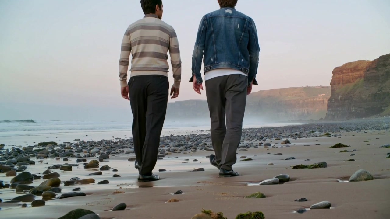 Walking pair heading toward cliffs on wet shore, camera pulling back, striped-sweater denim-jacket