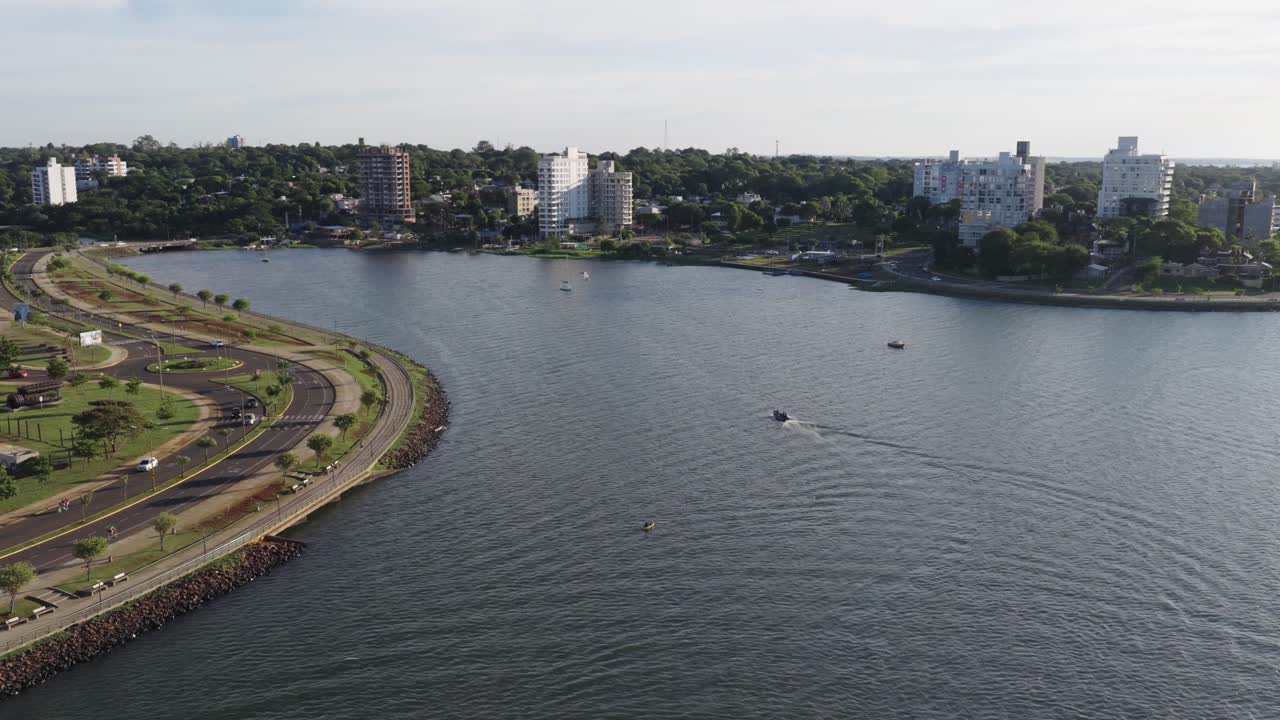 Aerial view of Posadas, Misiones, Argentina with Bahía El Brete, riverside avenue Teresa de Calcuta, waterfront buildings, green park areas, visible boats, and the city skyline, drone orbiting
