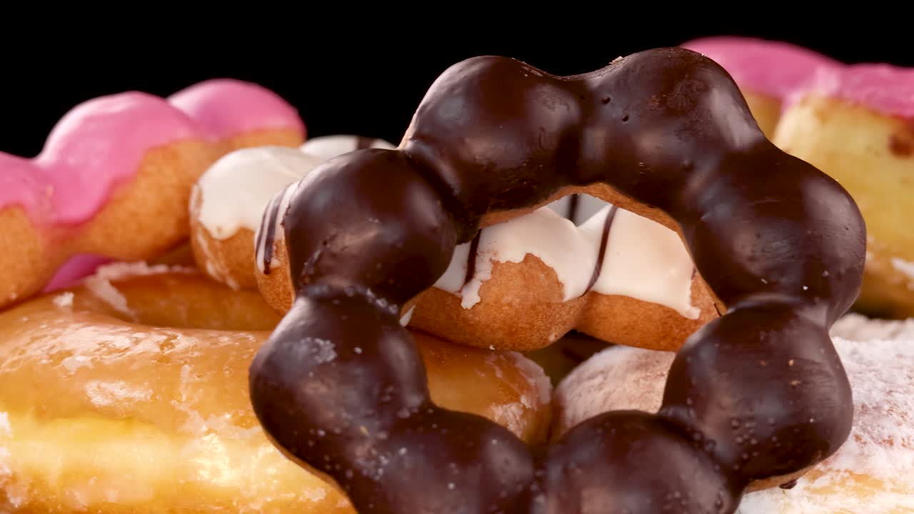 A colorful assortment of donuts, including heart-shaped and ring varieties with pink, chocolate, and white icing, rotates under bright studio lighting against a black background
