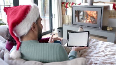 Pareja-Birracial-Con-Sombreros-De-Santa-Claus-Usando-Tableta-Con-Espacio-Para-Copiar-En-La-Pantalla-En-Casa,-Cámara-Lenta