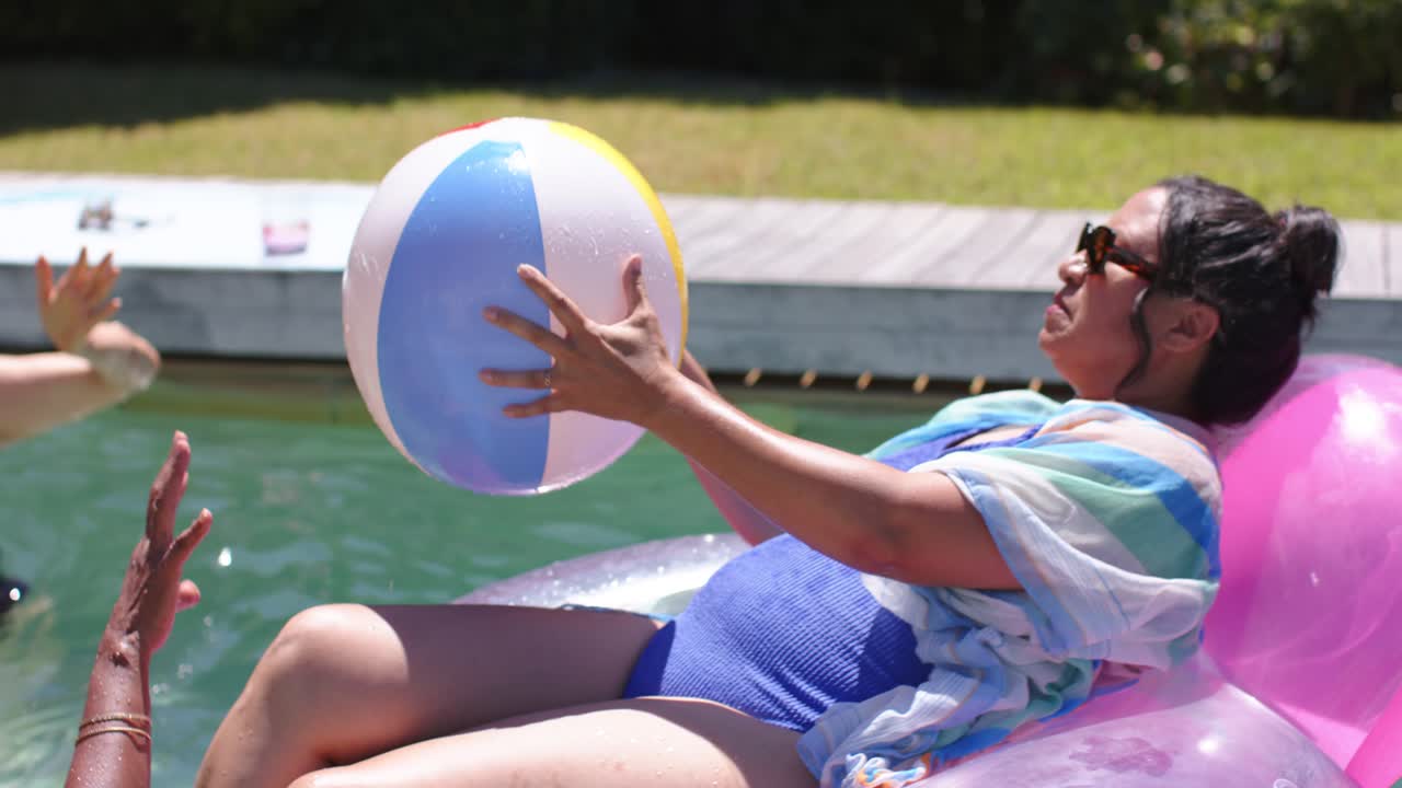 Raising ball, float woman and friends passing beach ball at backyard pool in swimsuits, cooling off