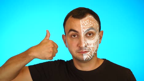 man with painted face showing thumb up and looking at camera against blue background