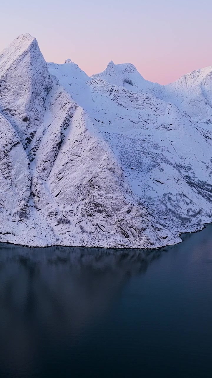 Atemberaubende vertikale Luftaufnahme von schneebedeckten Gipfeln, die sich in den ruhigen Fjordgewässern der Lofoten, Norwegen, während eines rosa Wintersonnenuntergangs spiegeln.
