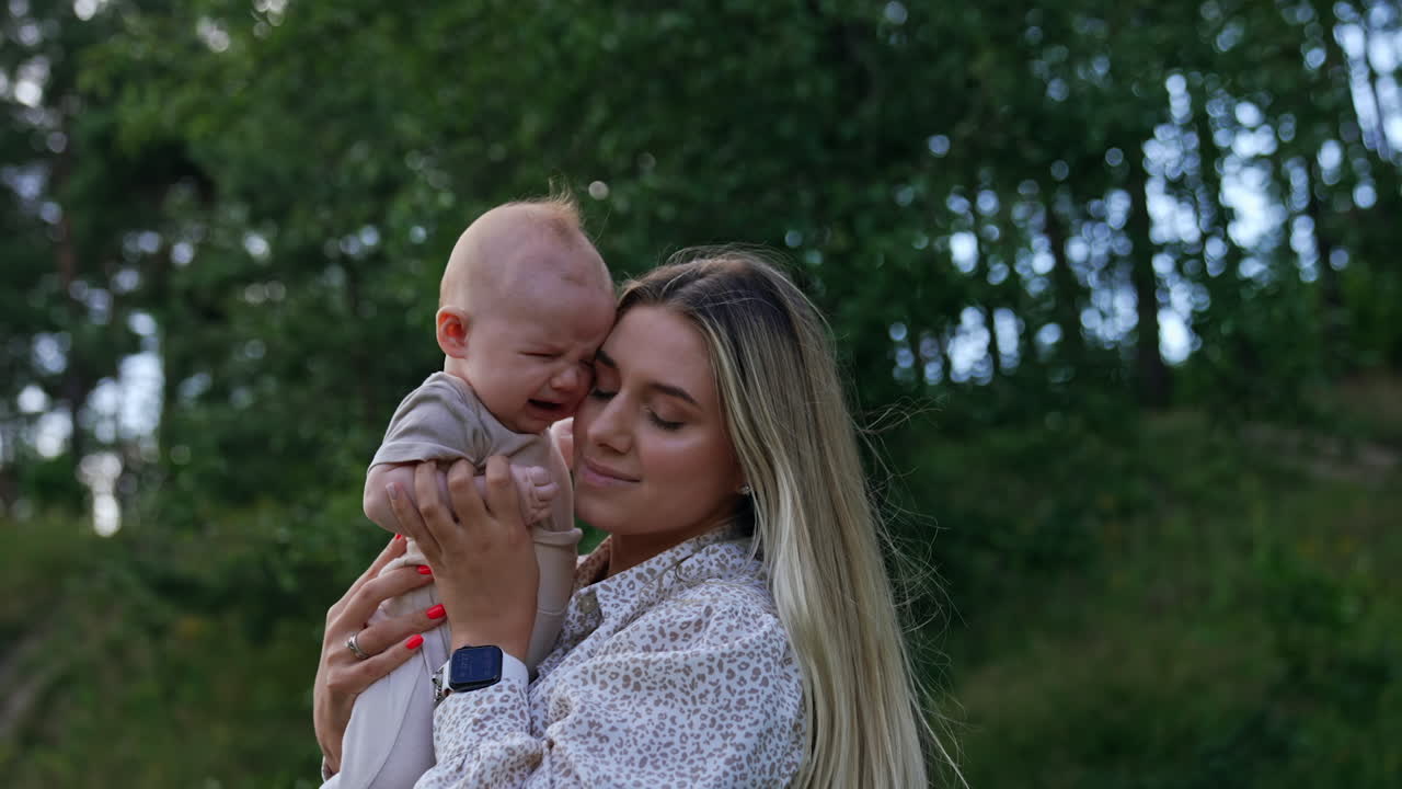 Little crying baby leaning to his mom's cheek. Adorable newborn crying bitterly as mom comforts him. Nature at backdrop.