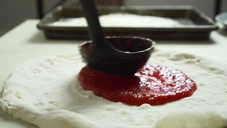 close up of chef while spreading tomato sauce on pizza dough with a ladle in traditional italian pizza's restaurant