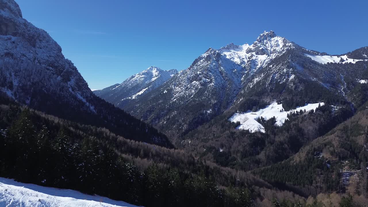 Drone fly passed snowy road towards pine forest with huge mountains with snow covered summits in background in Austria