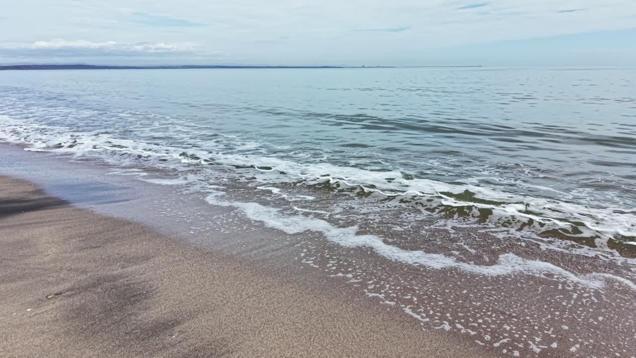 Bølger ruller ind på en sandstrand, mens skyer er spredt på himlen på en klar dag ved havet.