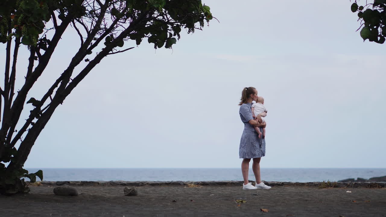 A picturesque portrait captures a mother's kiss shared with her daughter by the sea and beach. A content family on vacation, the mom envelops her toddler in a warm embrace