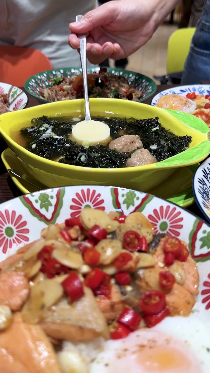 Una mano sirve tofu de una vibrante sopa de albóndigas de cerdo y algas en un tazón amarillo, rodeada de coloridos platos de arroz tailandeses bajo una cálida iluminación interior.
