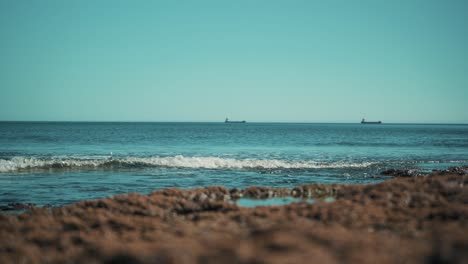 Costa-Oceánica-Plana,-Con-Barcos-De-Carga-Lejos-Y-Olas-Golpeando-Rocas-Suavemente-Al-Sol-4k