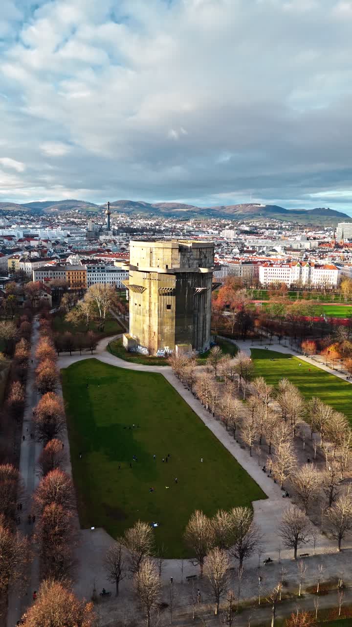 Luftfoto med drone, der panorerer til venstre mod Augarten-tårnet i Wien ved solnedgang om vinteren og viser skyskraberen, de omkringliggende bygninger, vinterlige skyer og varmt sollys, der reflekteres på hustagene