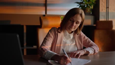 Concentrated-middle-aged-blonde-woman-in-a-pink-jacket-and-business-suit-sits-at-a-wooden-table-in-the-office-and-writes-down-her-ideas-on-paper-in-a-sunny-office-in-front-of-a-laptop