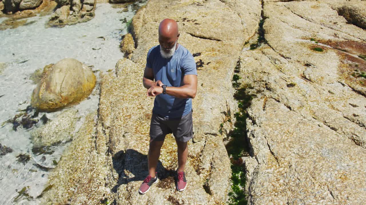 Senior african american man exercising using smartwatch on rocks by the sea