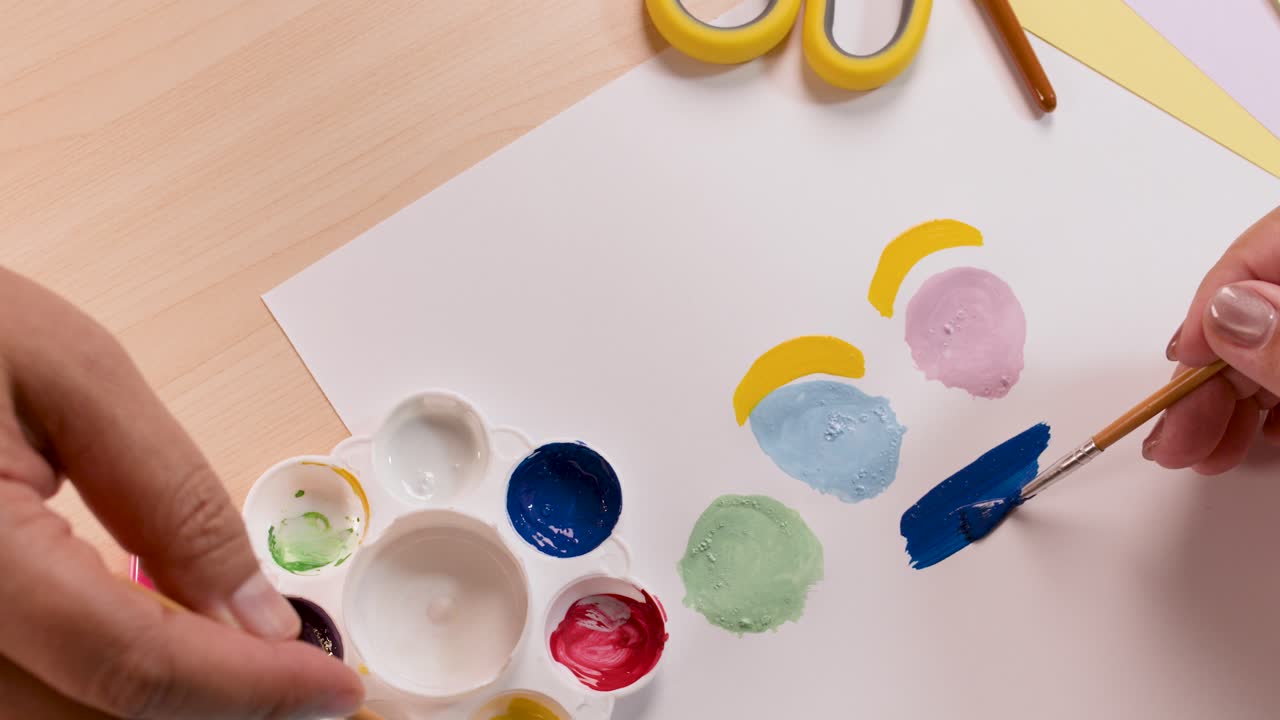 Child’s hands painting colorful shapes on paper with brush, bright lighting, overhead view