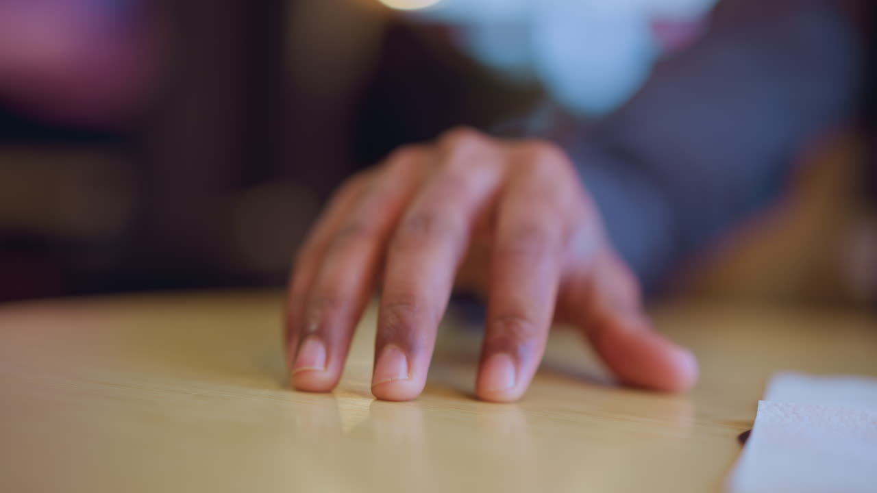 Close-up of young man's hand gently resting on table surface in soft indoor lighting, creating subtle emotion of hesitation, anticipation with blurred background suggesting quiet reflective environment