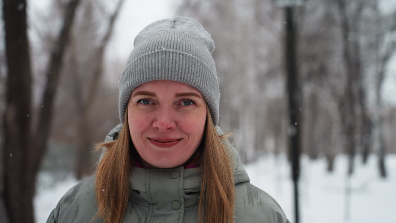 Donna con giacca invernale verde e berretto grigio in piedi su un sentiero innevato in un parco forestale, circondata da alberi spogli e neve che cade dolcemente, sorride con calma alla macchina fotografica durante una tranquilla giornata fredda.