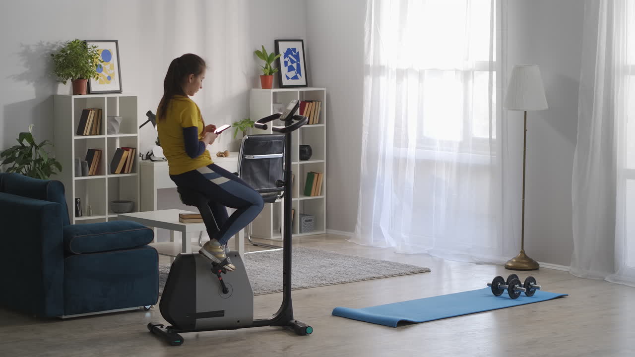 mujer está escribiendo y enviando mensajes por teléfono celular sentada en la bicicleta de ejercicio en la sala de estar de su apartamento descanso en el entrenamiento matutino estilo de vida deportivo y helathy