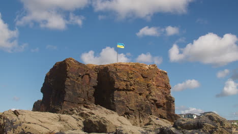 Ukrainian-Flag-On-Top-Of-Chapel-Rock-at-Perranporth-Beach,-Cornwall,-UK---wide-shot