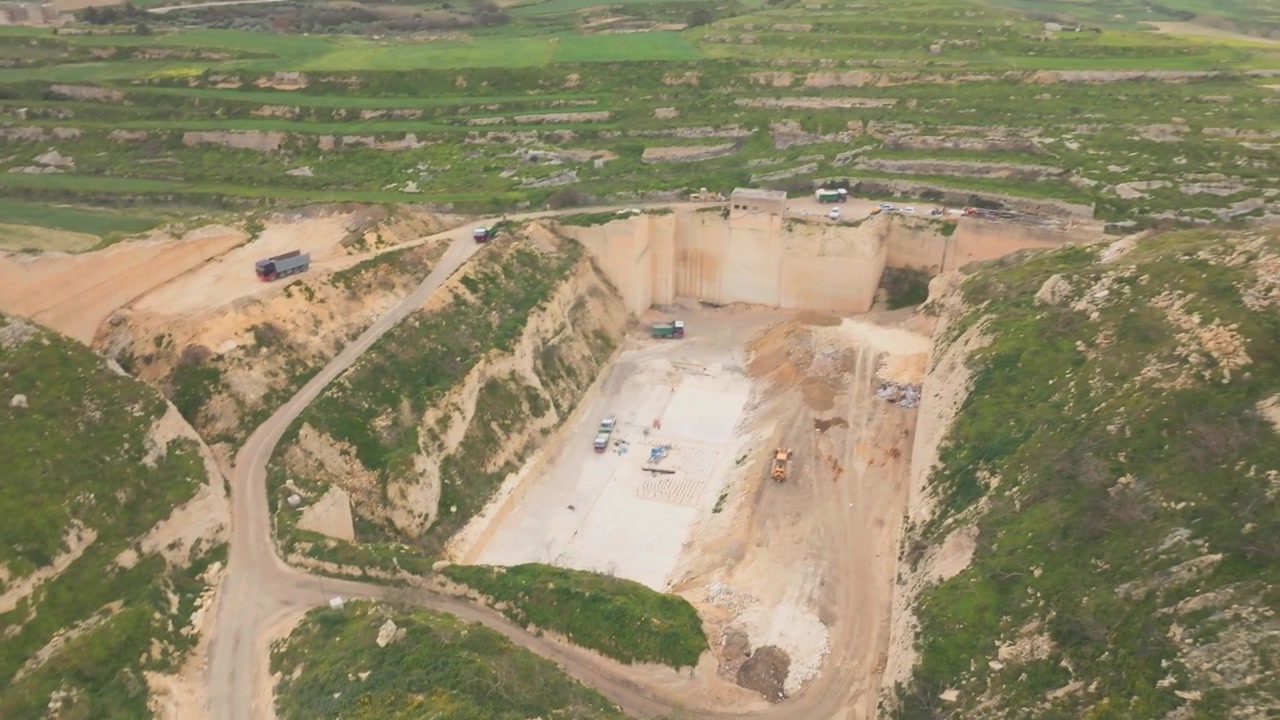 Una vista aerea di una grande cava di pietra circondata da colline verdi terrazzate, con camion e attrezzature da costruzione che si muovono lungo strade sterrate e il fondo della cava.