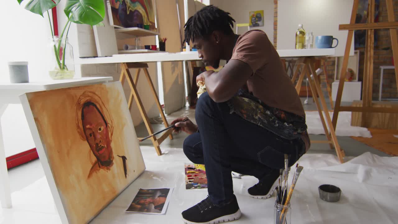 vista lateral de un artista masculino afroamericano con un delantal pintando en lienzo en un estudio de arte